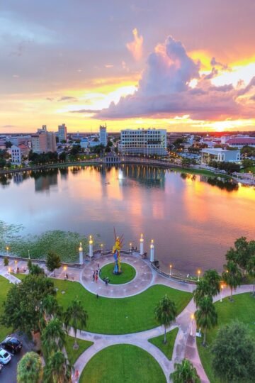 Aerial view of Downtown Lakeland, Florida at sunset, featuring the glowing skyline reflecting on Lake Mirror. The foreground shows the Promenade with green lawns, palm trees, and the 'Tribute to the American Spirit' statue in a circular plaza.