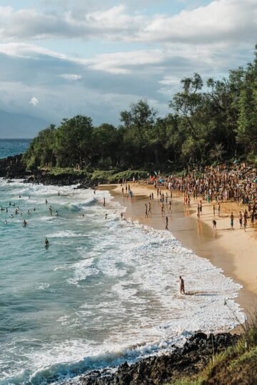 Crowds enjoying the ocean and sandy shore at Little Beach, a popular nudist beach in Hawaii surrounded by lush greenery and volcanic rocks.